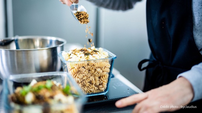 Préparation minutieuse : l'ajout de saveurs à votre plat Cookingo Une personne ajoute des graines (tournesol, courge, etc.) à une portion de pâtes déjà préparée dans un récipient transparent, dans un contexte de cuisine Cookingo.