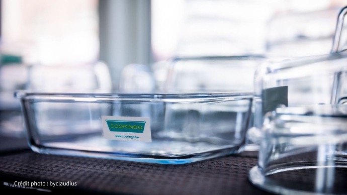 Vos repas Cookingo conservés avec soin dans nos boîtes Un récipient rectangulaire en verre transparent avec un autocollant du logo Cookingo est mis en évidence sur une surface sombre, entouré d'autres contenants en verre flous.