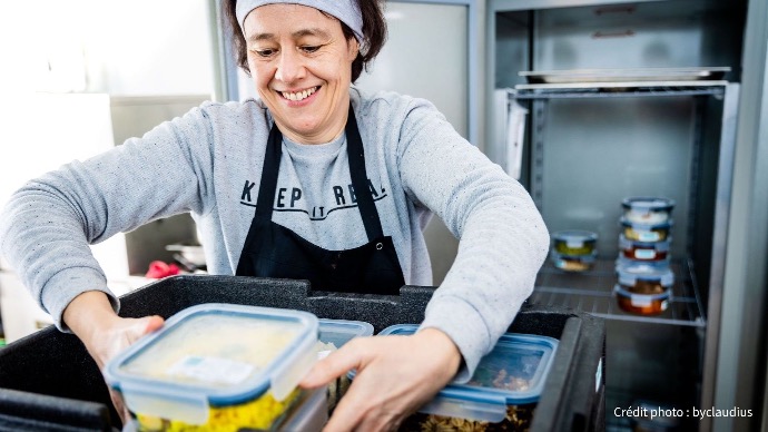 Préparation des commandes Cookingo : l'assemblage de vos repas Une femme souriante, portant un tablier noir, organise des boîtes de repas préparés dans un grand bac noir, dans un environnement de cuisine professionnelle.